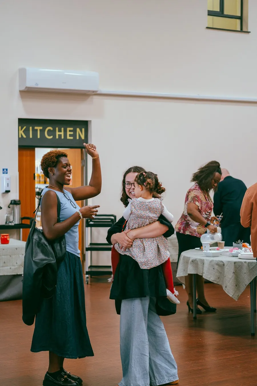 A woman with a toddler in her arms talking with friends in the church hall beside the kitchen